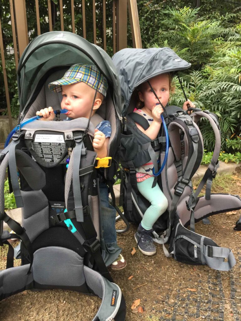 two toddlers sit in their backpack carriers for hiking. Both children have a sun canopy and are drinking from their hydration systems.