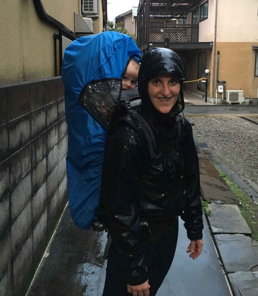 A mom stands in the rain with her toddler in Kyoto, Japan. She keeps her son dry with a rain cover on her hiking backpack for toddlers.