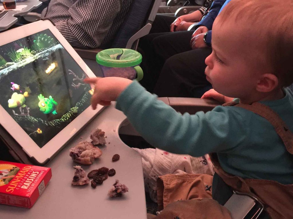 a busy toddler playing with ipad and eating snacks on a long flight with his family