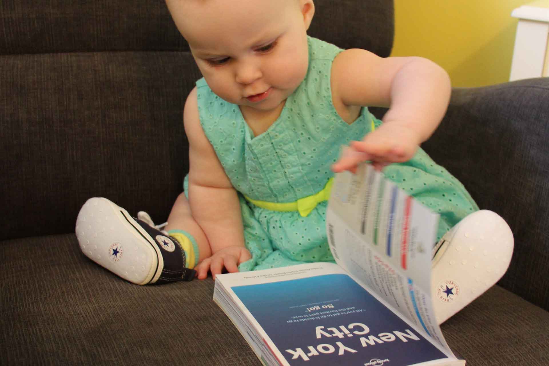 A very cute baby in a nice dress looks at a New York City Lonely Planet travel guide .