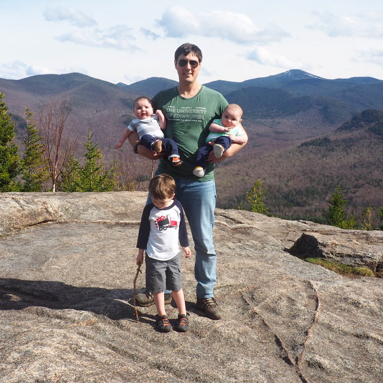 a proud father poses for a picture with his toddler and twins on a family vacation.