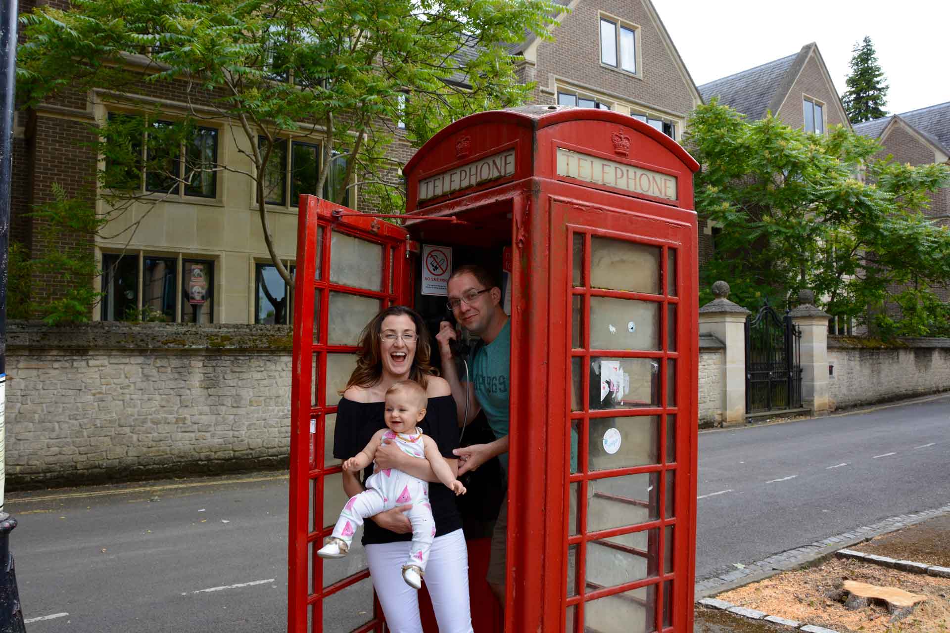 a family poses for a fun picture in an iconic London phone booth with their baby.
