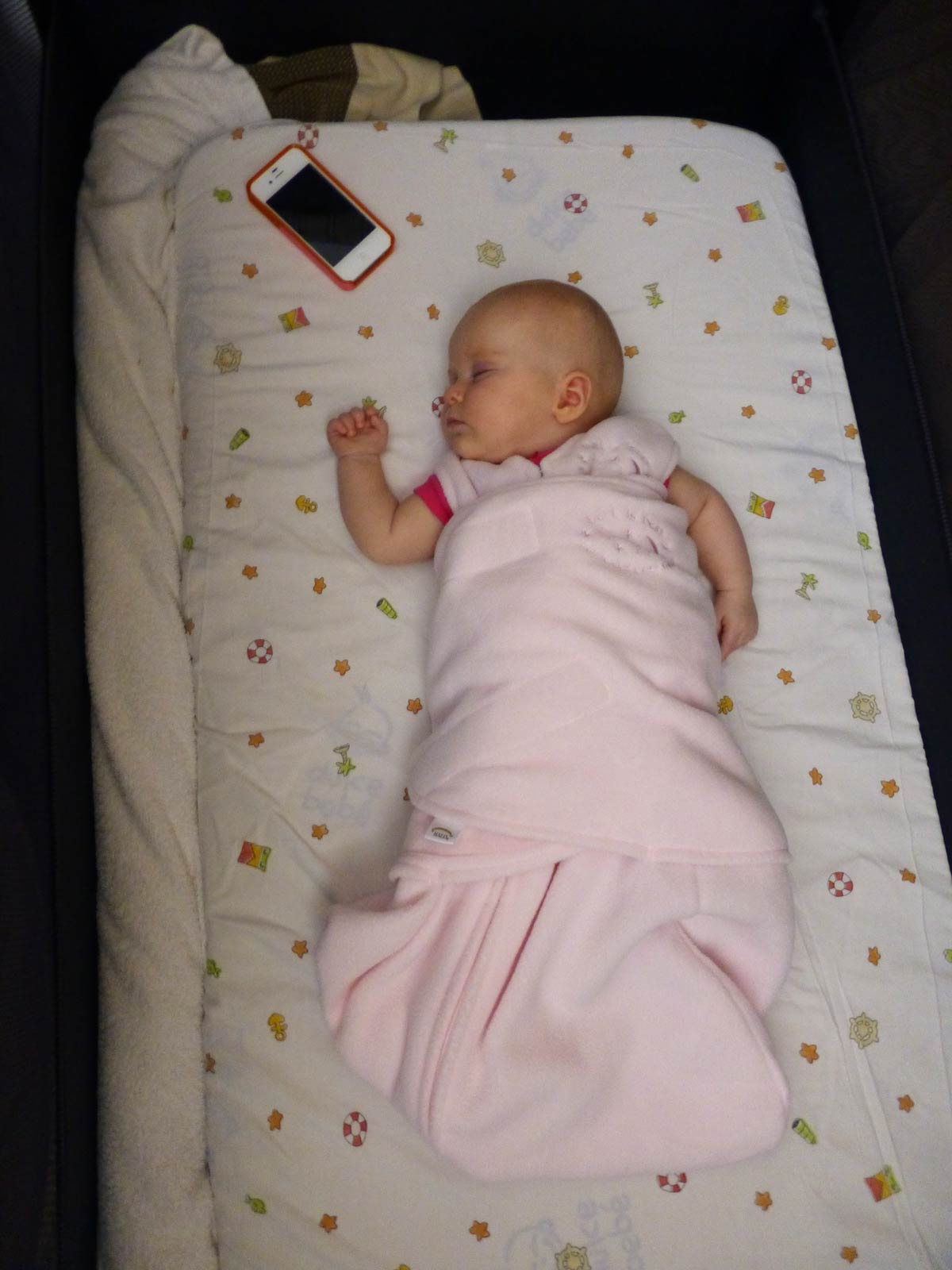 A baby from the BabyCanTravel.com family, sleeps in a Halo Swaddle Sleepsack with an old iPhone providing white noise.