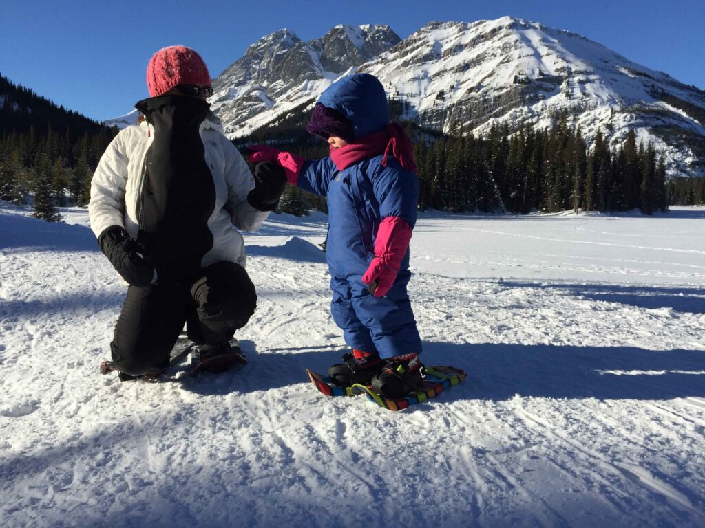 a mom carrying an infant inside her winter jacket goes snowshoeing in winter in Canada with her toddler.