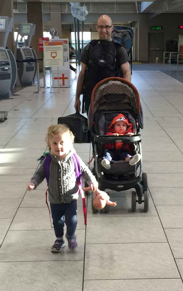 A Dad pushes his infant in a stroller while his toddler runs ahead at the airport ahead of a family vacation