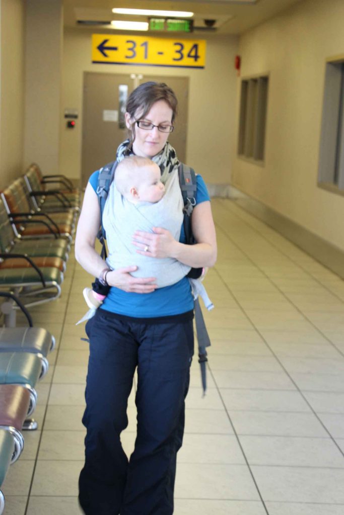 A mother carries her infant in a baby carrier walking through the airport prior to her flight