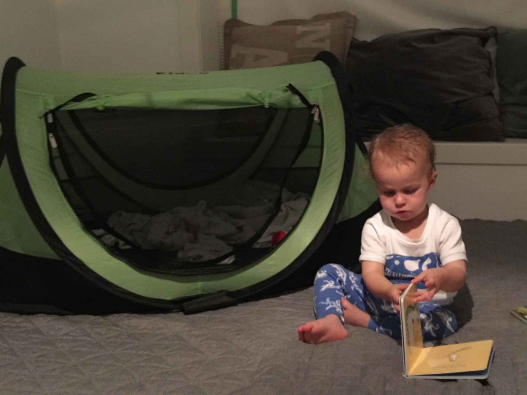 a toddler reading a book at bedtime near his toddler travel bed. In order to make the room dark, we used painters tape and cushions to help close the curtains to try and remove the extra light coming into the room