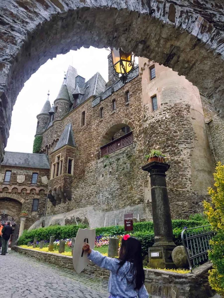 a toddler with a play shield stands in front of a castle archway during a family trip to Germany
