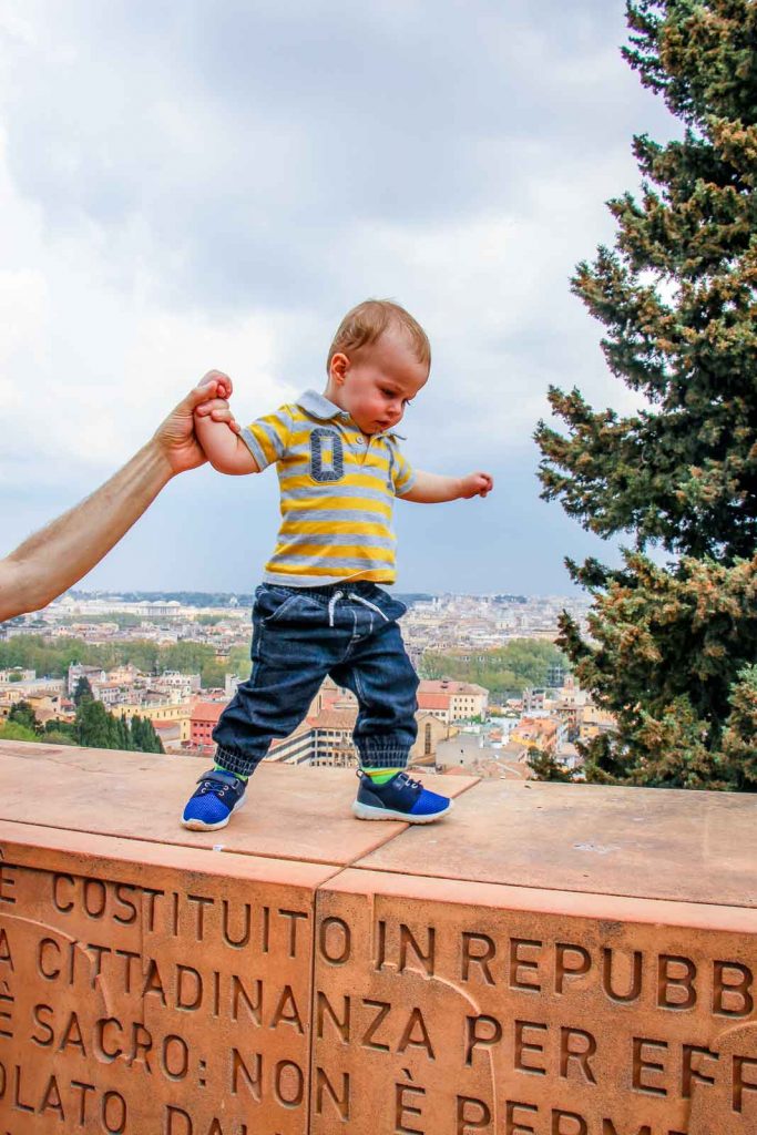 a toddler walks atop a stone wall overlooking Rome, while on a family vacation to Italy