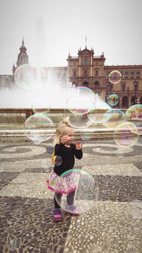 a toddler is surrounded by huge bubbles in a beautiful square during a family trip to Spain