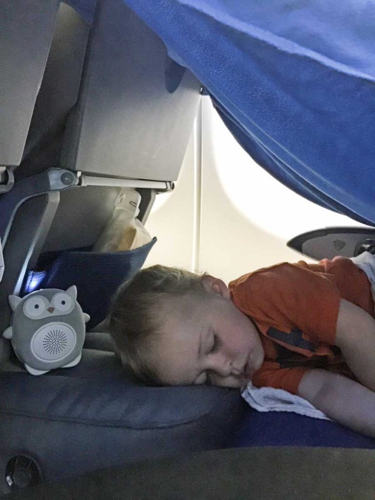 toddler sleeping on airplane cushion with white noise machine also sitting on cushion.