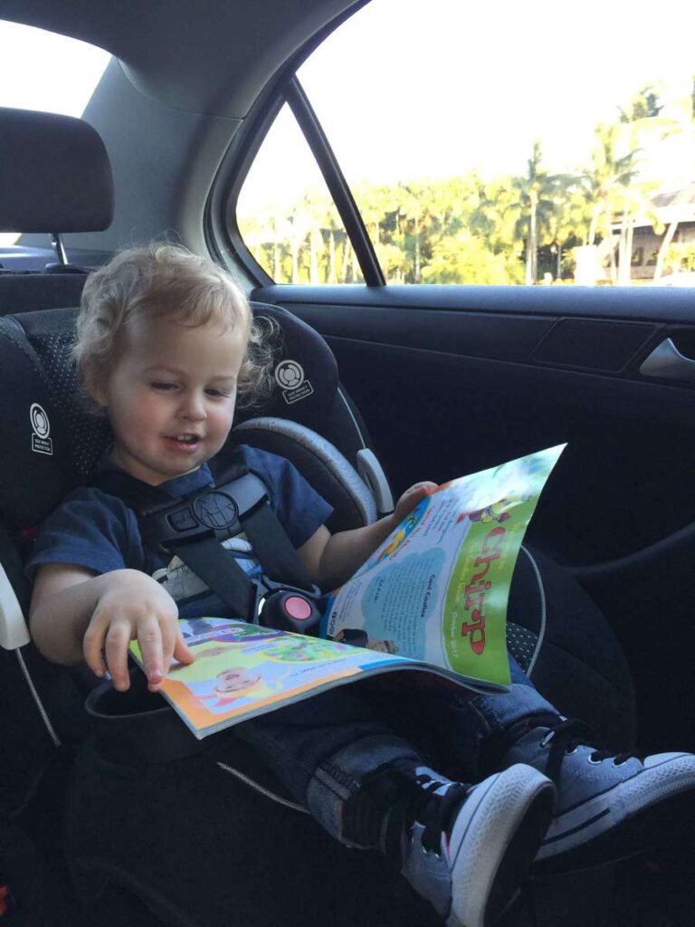 a toddler sitting in a travel car seat smiles while reading a magazine