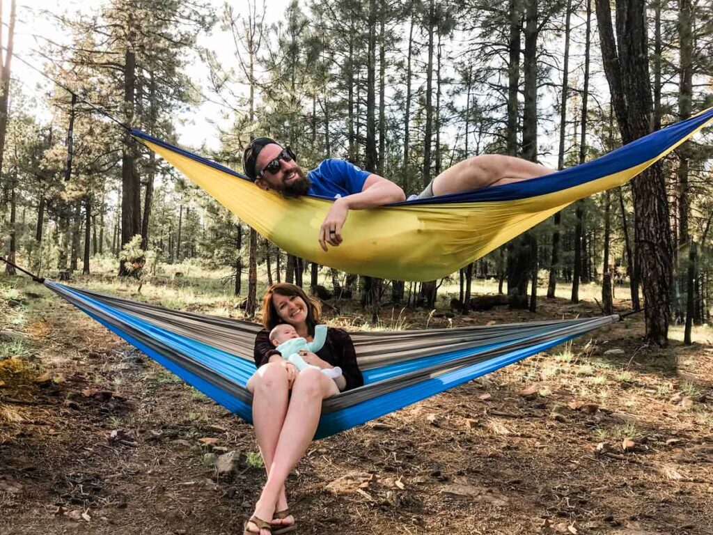 two parents lie on hammocks in the forest while tent camping with a baby