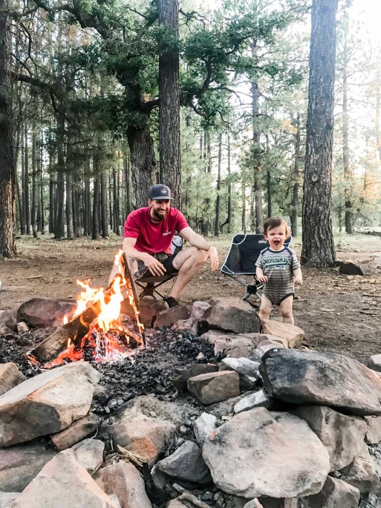 an excited baby enjoys standing by the campfire while camping with his dad in the forest