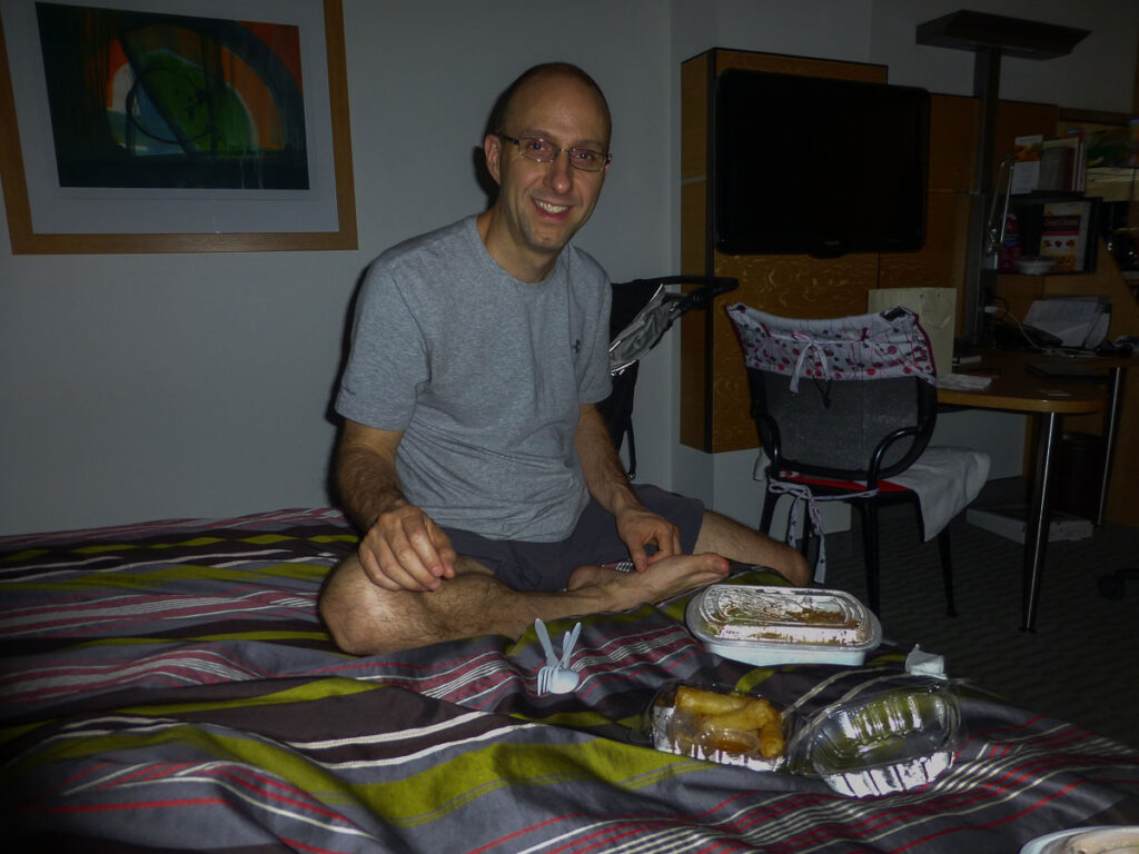 A man prepares to eat dinner in a dark hotel room in New York City, trying to not wake his sleeping baby a few feet away.