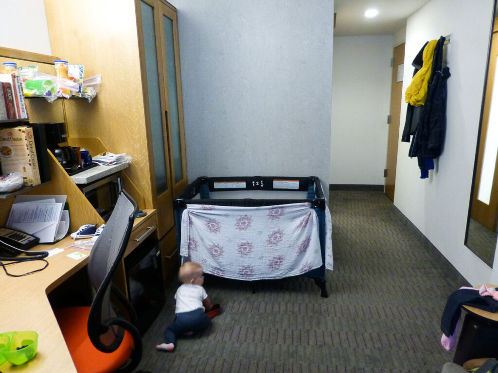 a baby crawls on the floor of her parents' NYC hotel room - the pack and play has a blanket on one side to prevent the baby from seeing her parents while sleeping.