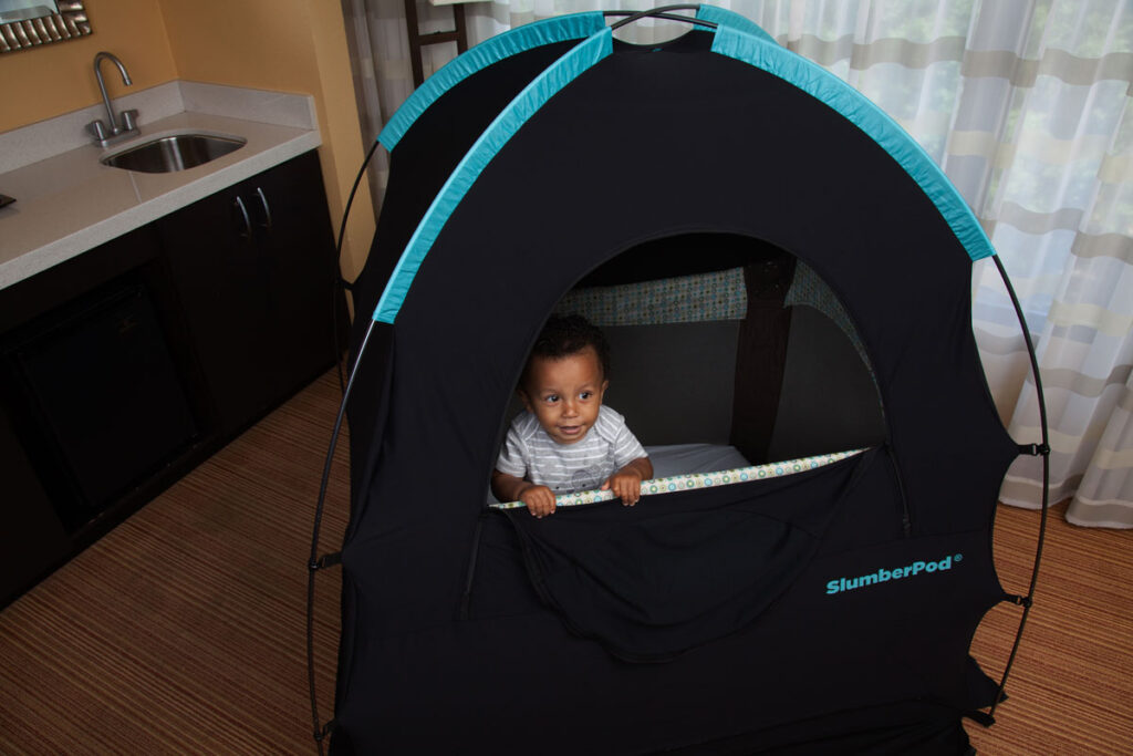 a toddler boy stands in a pack n play inside a SlumberPod privacy sleep nook