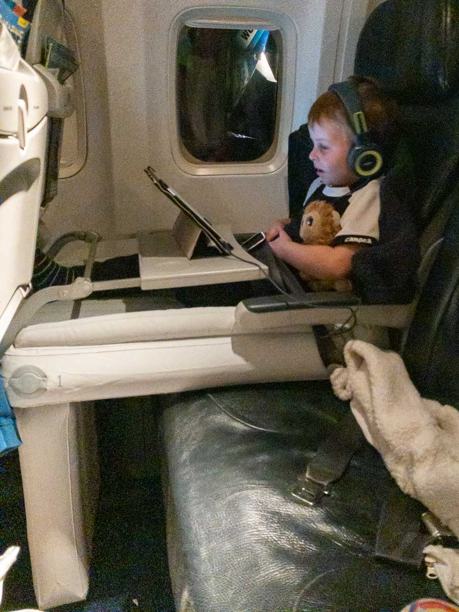 a toddler from BabyCanTravel.com gets comfortable on his Flyaway Kids bed on a flight while watching a movie on an ipad holding his stuffy.