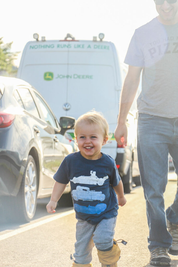 image of toddler walking beside cars on road with father behind him