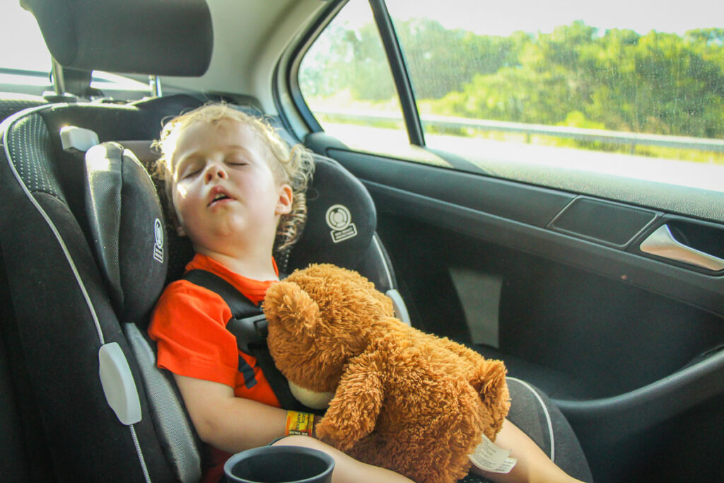 a child sleeps in his car seat holding a brown teddy bear during a long car trip with his family.