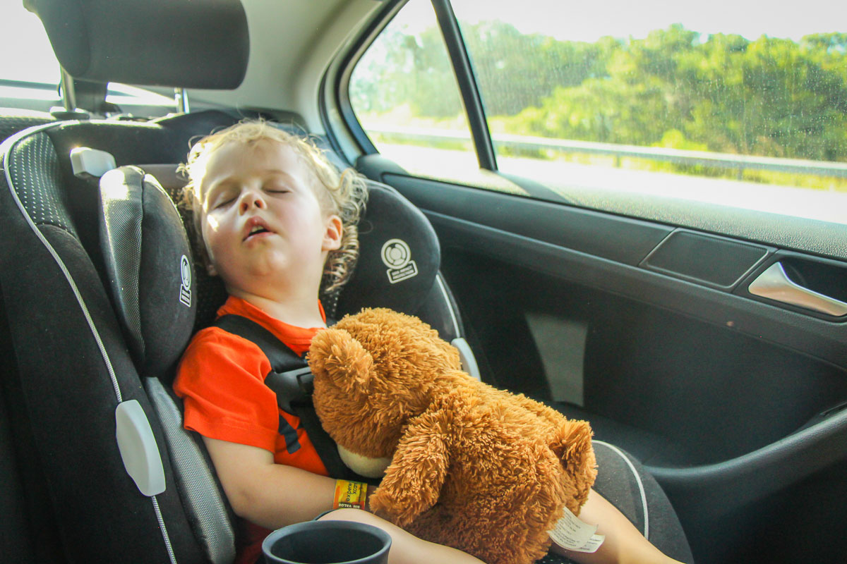 a sleeping toddler in a car seat during a family road trip vacation. The toddler is holding his favorite stuffy.