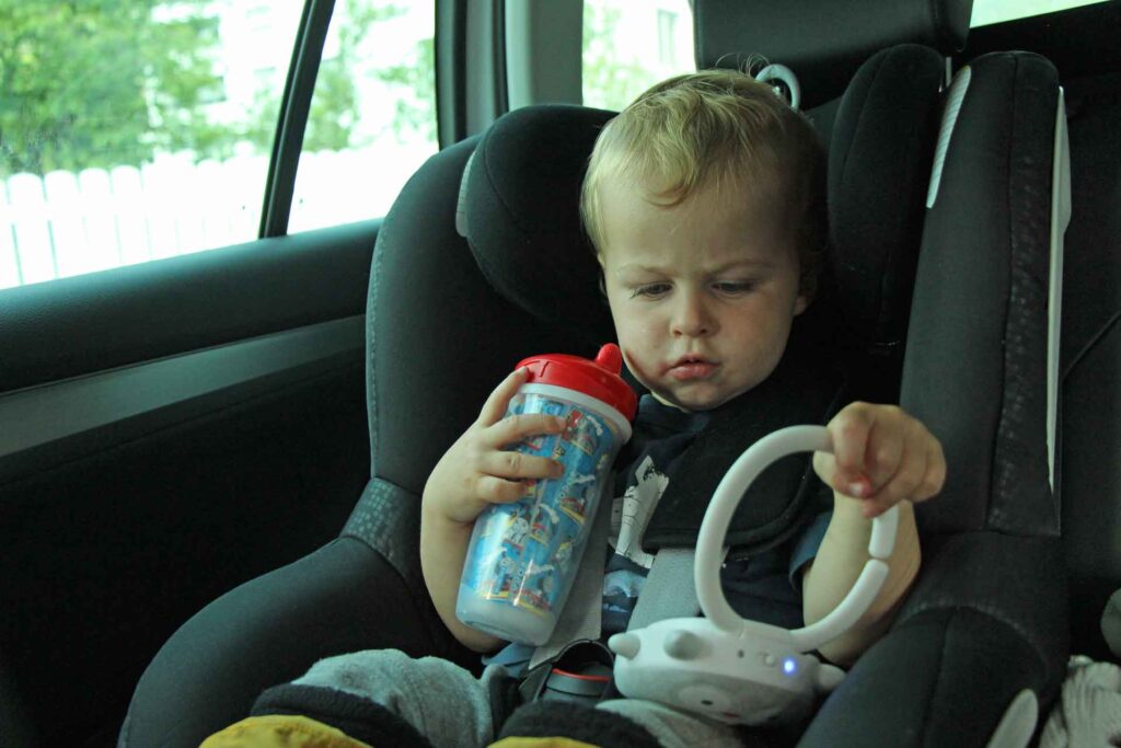 A toddler in a car seat holds a toddler travel cup and a white noise machine