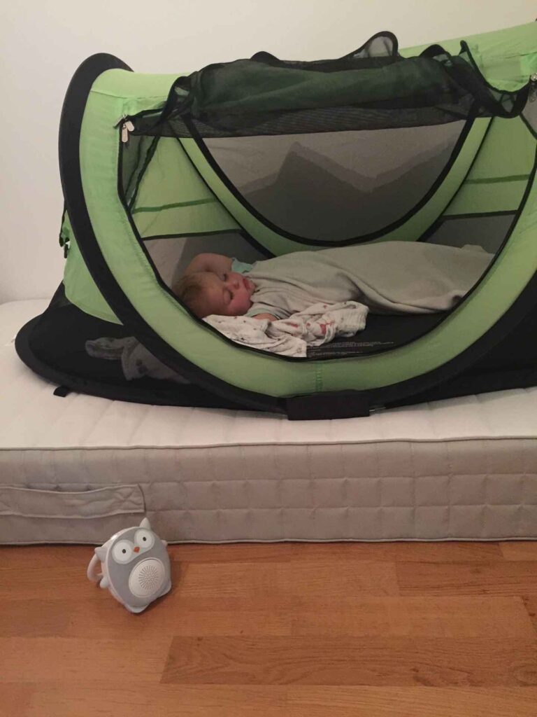 A toddler sleeps in a PeaPod Plus travel bed while using a SoundBub white noise machine