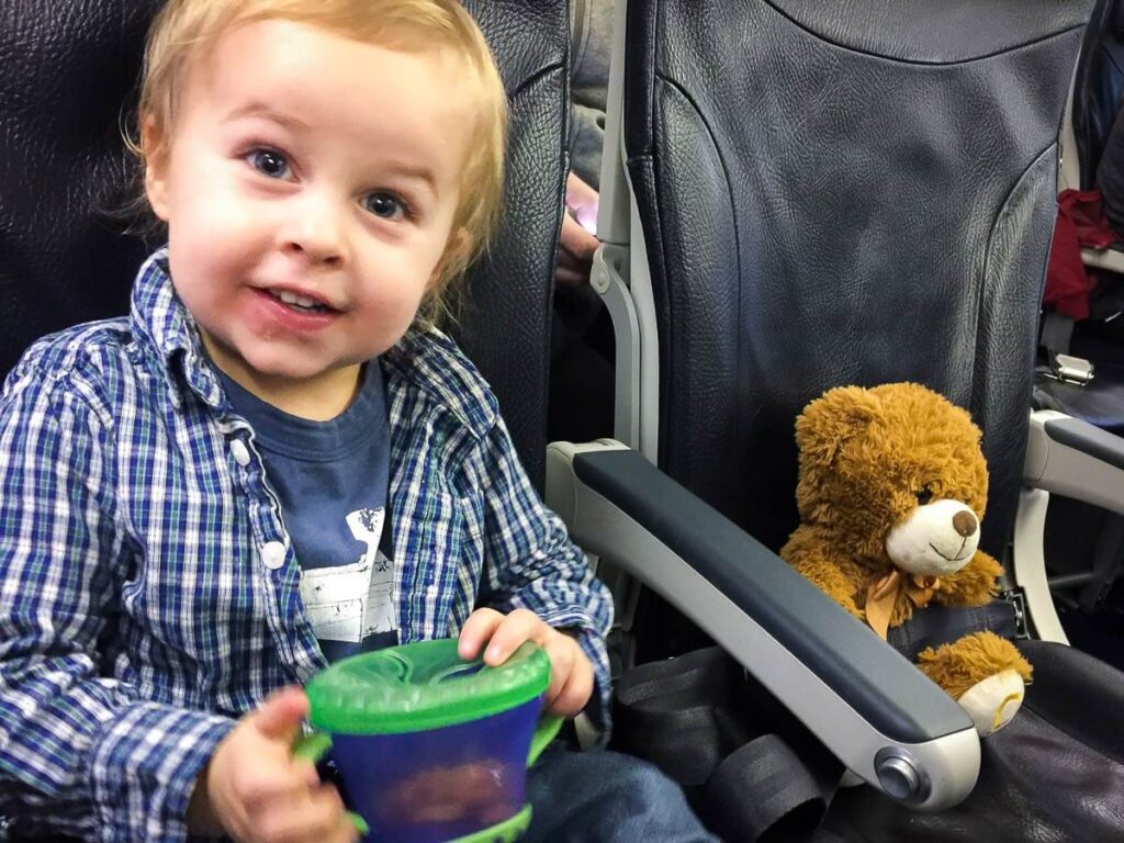 A happy toddler has a snack on a plane, while his stuffy sits next to him.