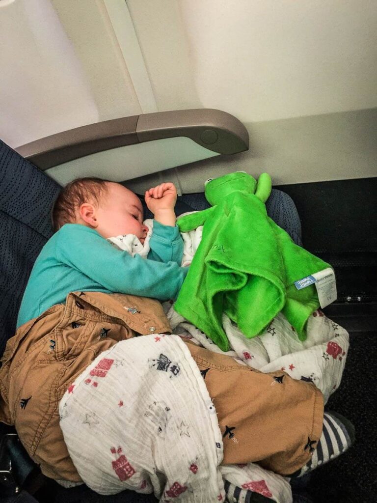 a toddler from the BabyCanTravel.com family sleeps on an airplane with his stuffy and an Aiden & Anais swaddle blanket.