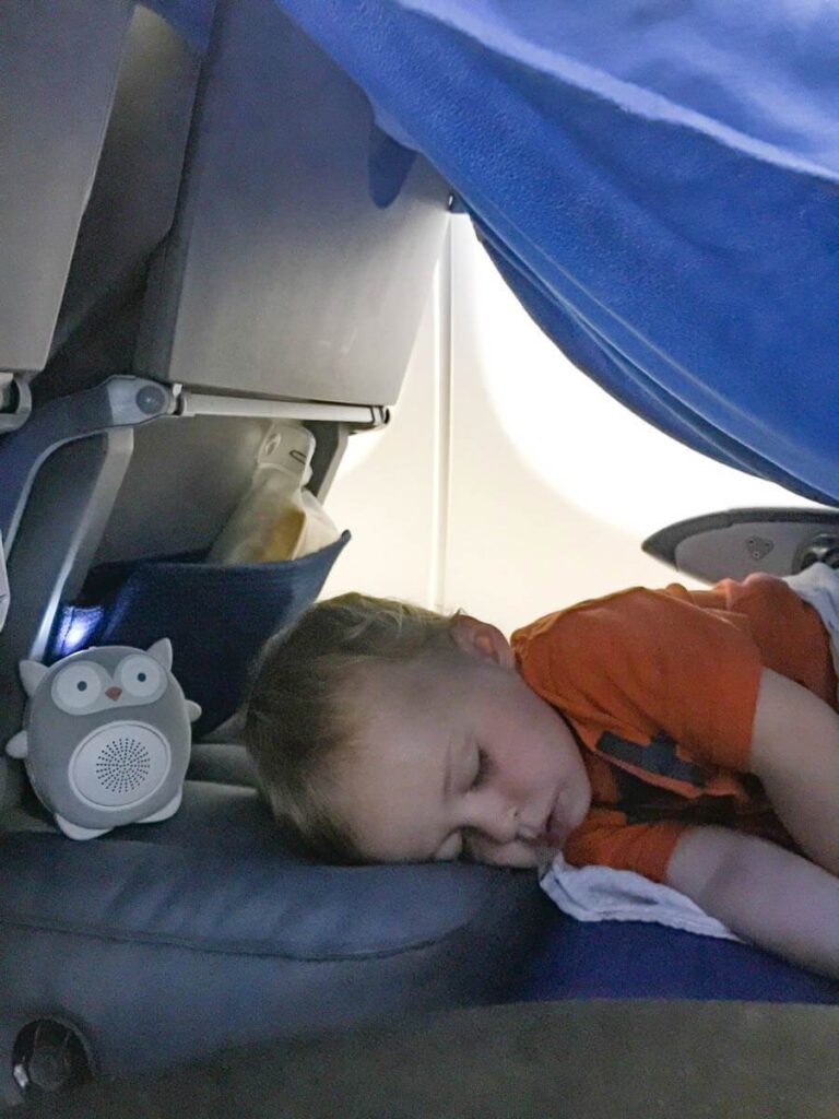 a toddler sleeping on airplane on inflatable airplane travel bed with a white noise machine and a blanket for a tent.