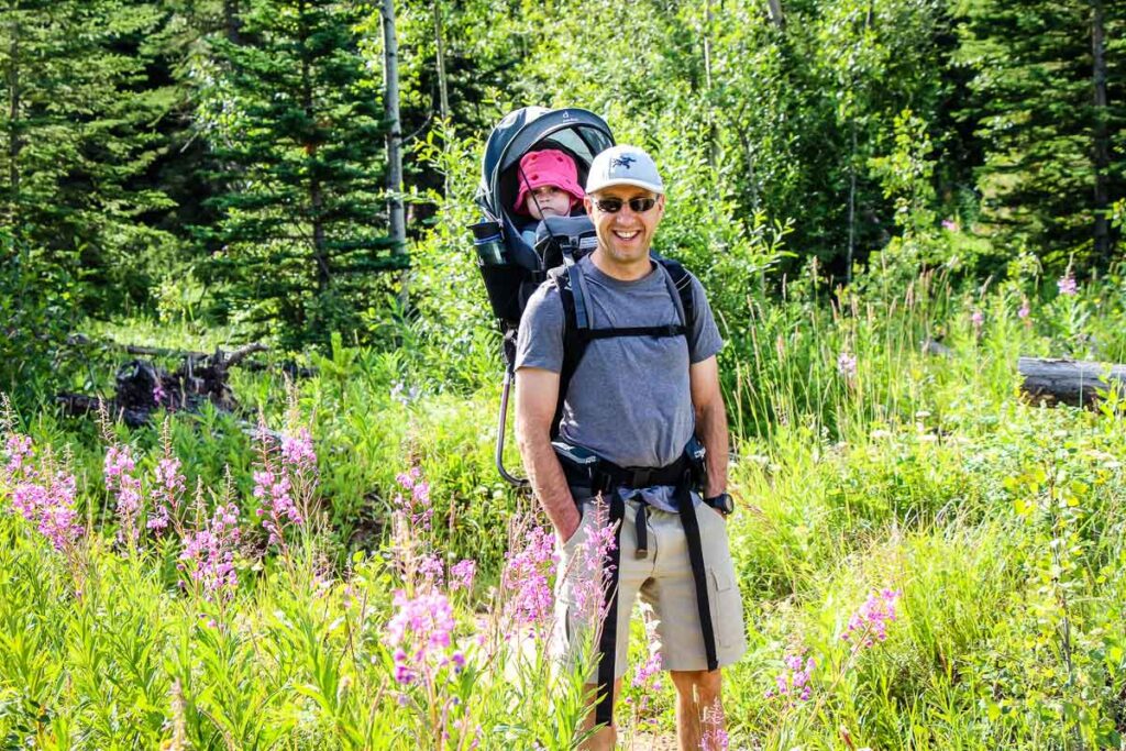 a father carries his toddler daughter in a child backpack carrier for hiking.
