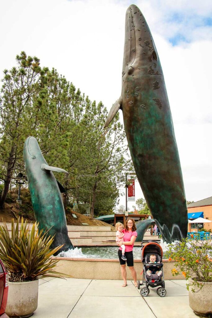 a mom, toddler and baby pose in front of the humpback whale sculpture at Birch Aquarium Scripps - one of the most fun things to do in san diego with toddlers.
