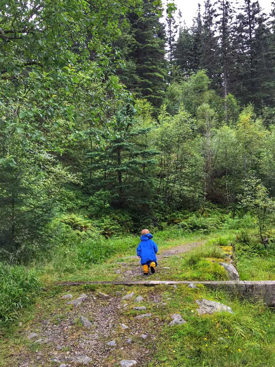 a toddler, from the Baby Can Travel family,  wears a rain suit and rain boots while hiking in the rain with his parents.