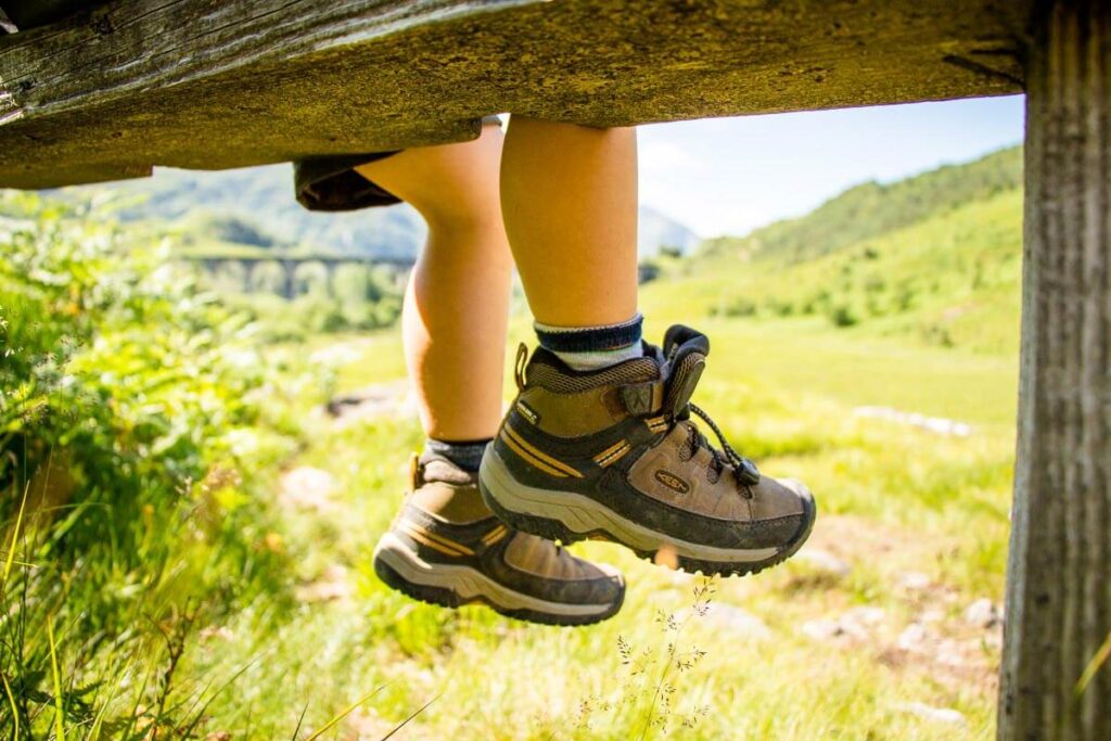 a toddler wearing Keen hiking boots rests on a wooden bridge with his feet dangling over the edge.