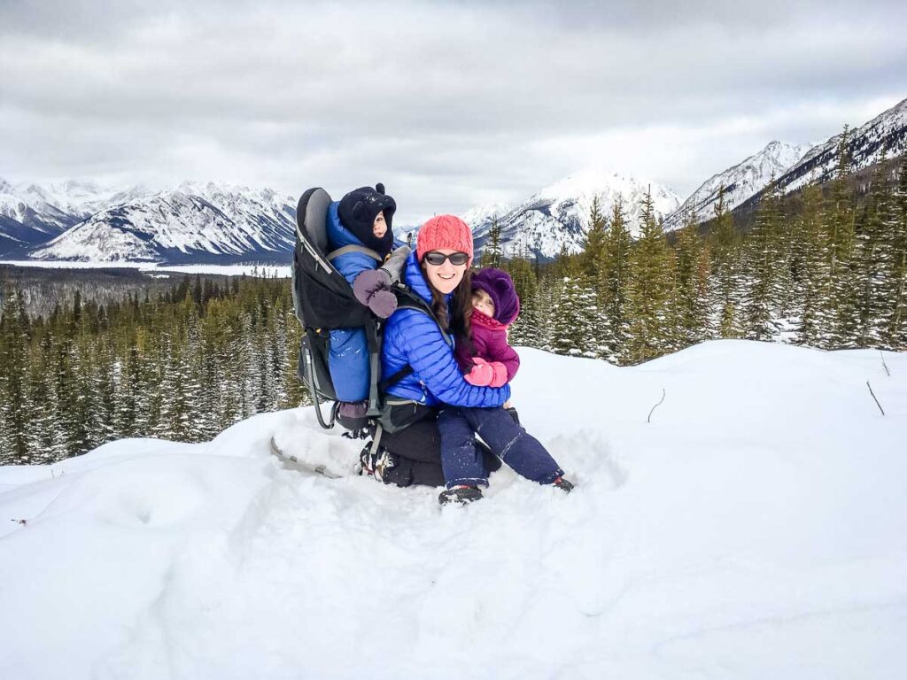 a mom hugs her preschooler and carries her toddler in a hiking backpack carrier on a family hike in winter in Kananaskis, Canada.