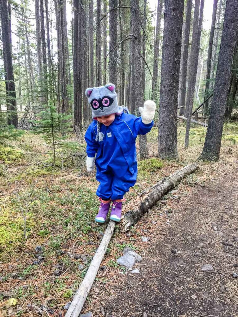 a toddler, from BabyCanTravel.com, wears a rain suit while balancing on a fallen tree trunk while camping with her family.