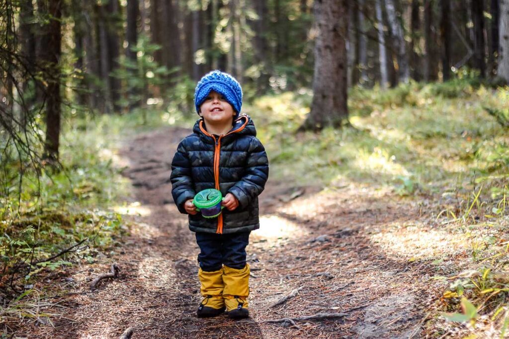 a hiking toddler, from the BabyCanTravel.com family, holds on to a spill proof snack cup in the forest.