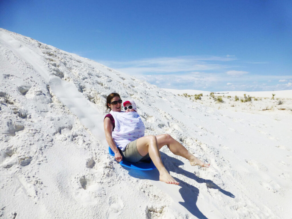 A mom enjoys sliding down a dune while visiting White Sands National Park with a baby