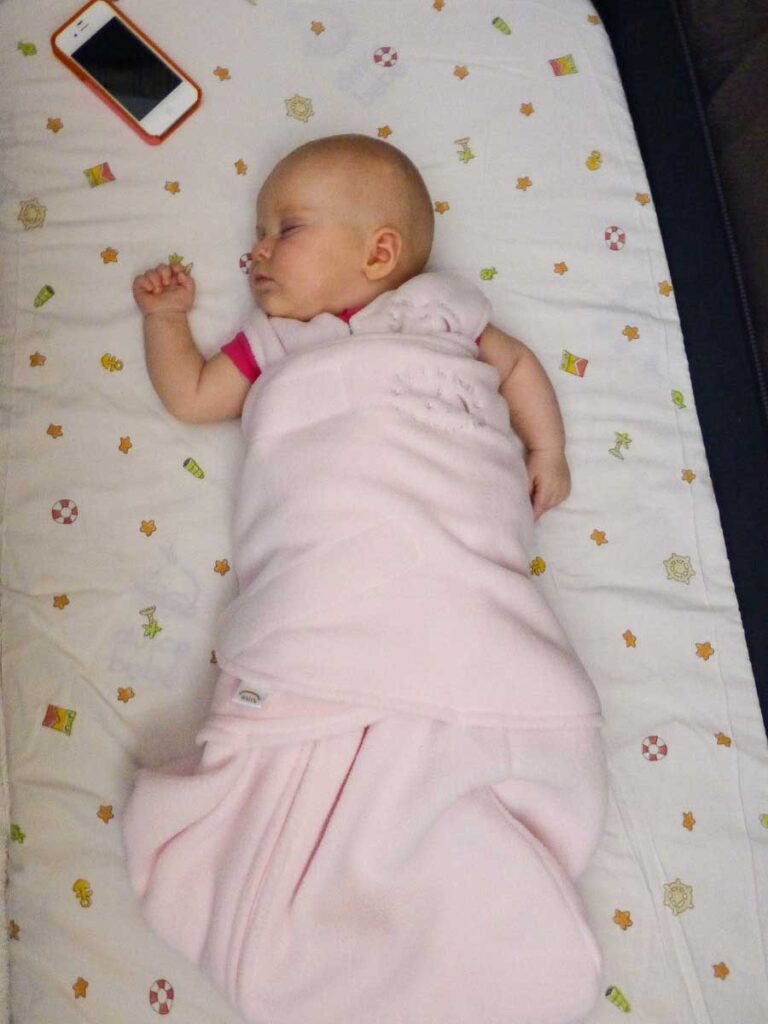 a baby girl sleeps in a hotel crib wearing a sleep sack and listening to a white noise app on a nearby iPhone.
