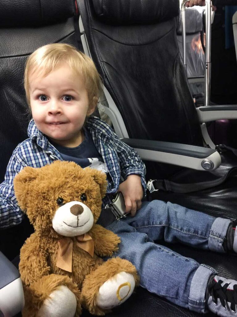 a toddler from Baby Can Travel sits nicely with his brown teddy bear while on a flight for his family vacation.