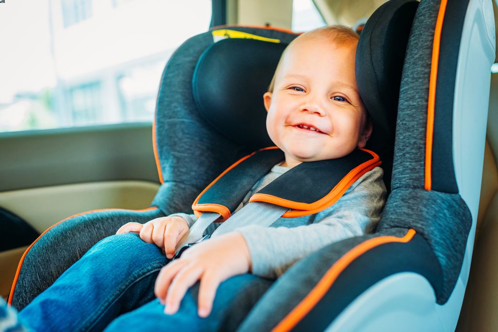 a baby smiles while sitting in a front facing car seat in the back seat of a car