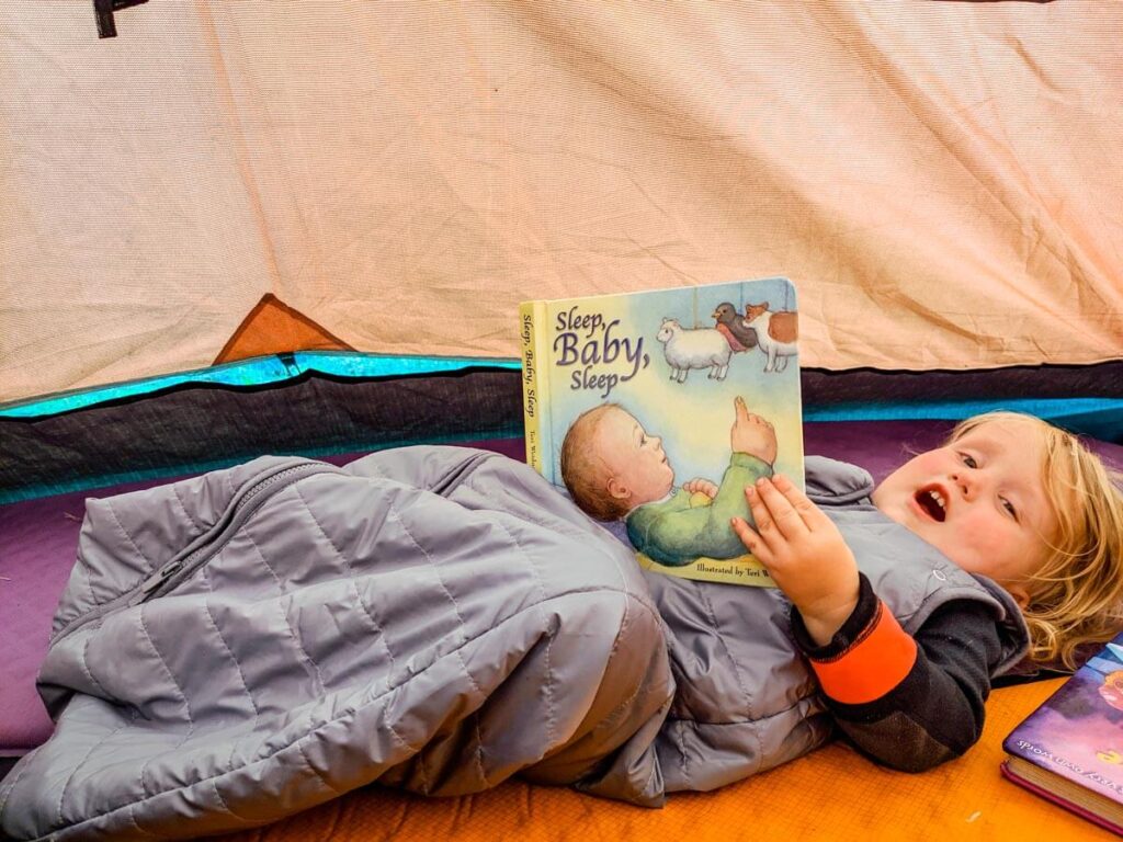 Toddler reading book and wearing Baby Deedee toddler sleeping bag for camping.