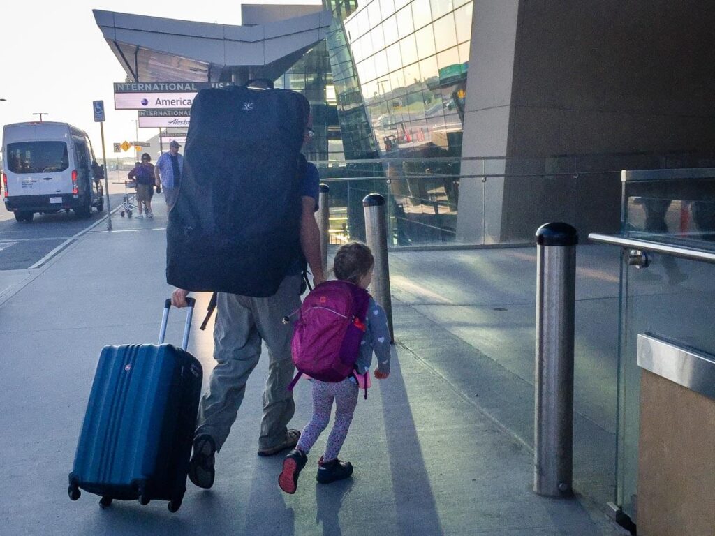 father and toddler walking into airport carrying JL Childress car seat bag for travel.