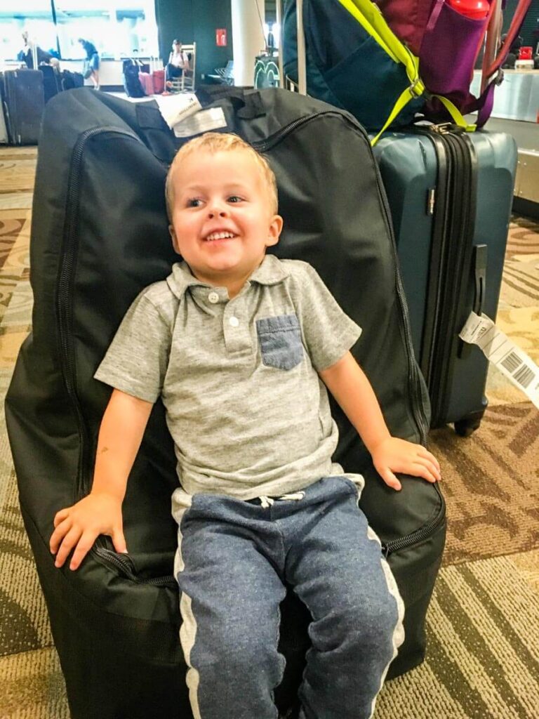 a laughing toddler in airport sitting on car seat in car seat bag for flying.