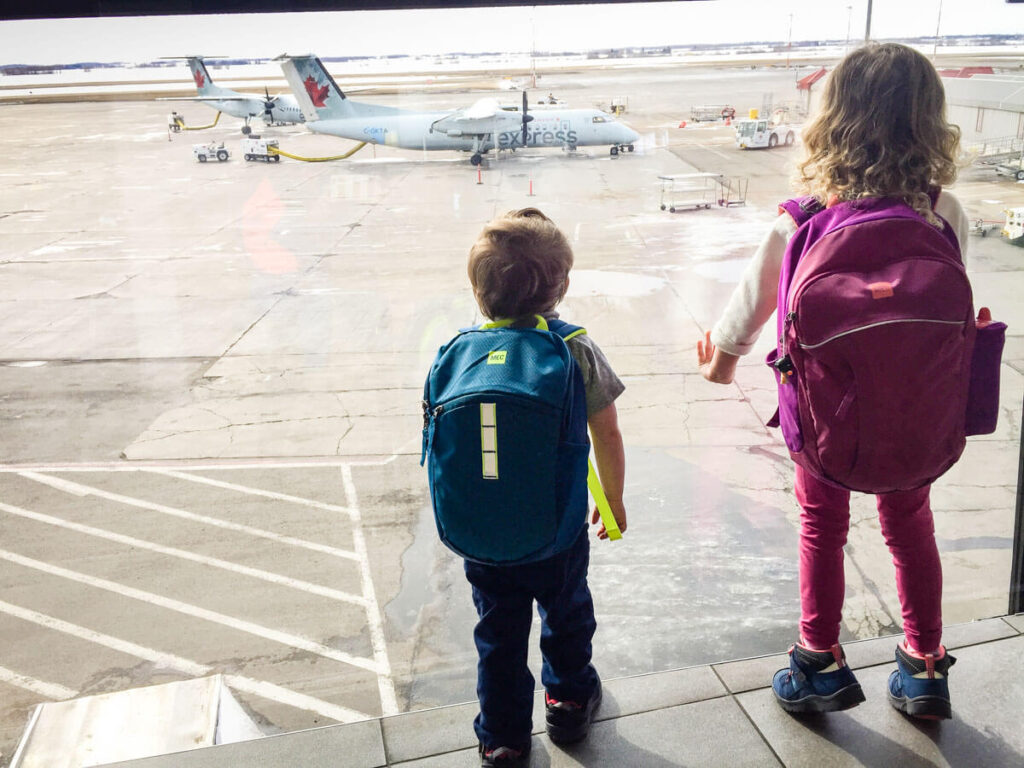 two toddlers wear their own toddler backpacks for airplane as they wait for board a flight at the airport.