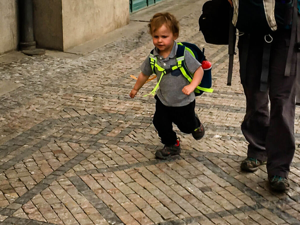 a small child wearing a toddler travel backpack runs on a cobblestone street on a family trip to Europe.