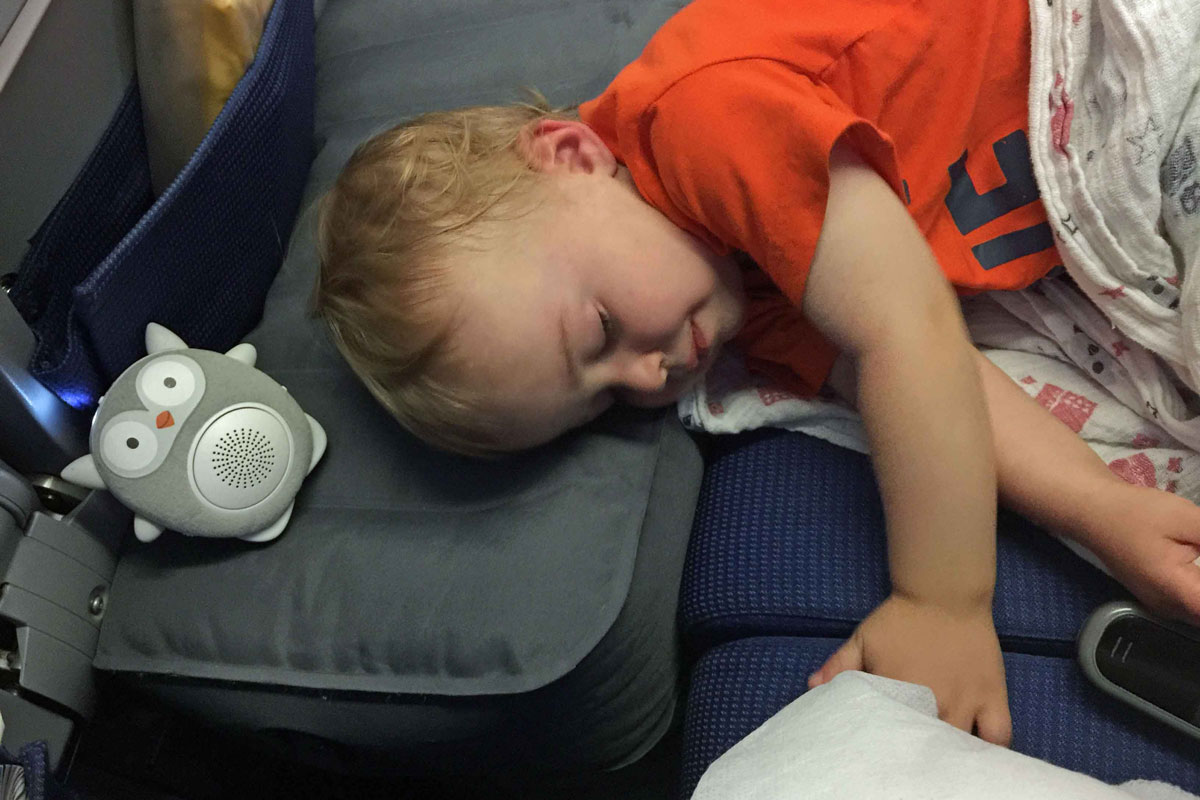 A toddler sleeps on an airplane using an Original Fly Tot inflatable airplane cushion.