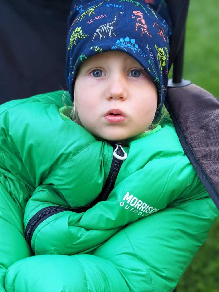 A toddler sitting on a chair in Big Mo 20 Sleeping Bag while on a family camping trip.