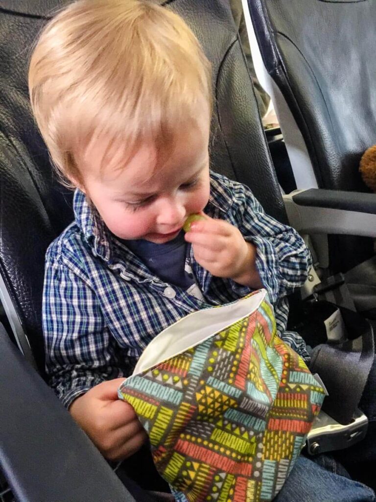 a toddler on an airplane eats grapes out of a toddler snack container.