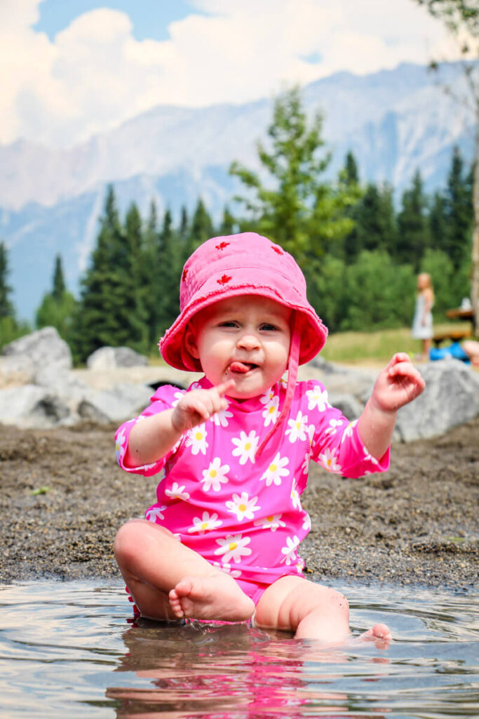 Baby sitting in lake with mountains in background wearing one of the best baby girl sun hats